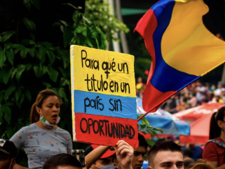 People protesting on the background. In close-up a sing painted in Colombian flag colours: yellow, blue and red. Which says "Why a degree in a country without opportunities”