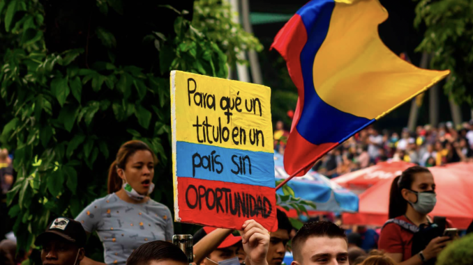 People protesting on the background. In close-up a sing painted in Colombian flag colours: yellow, blue and red. Which says "Why a degree in a country without opportunities”
