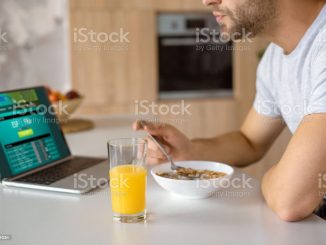 cropped image of man eating flakes with milk at kitchen table with fresh juice in glass and laptop with sportsbet on screen