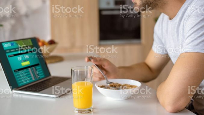 cropped image of man eating flakes with milk at kitchen table with fresh juice in glass and laptop with sportsbet on screen