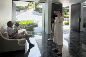 Photo showing an open house event led by real estate agent Vincent Bai. In the foreground, a woman in a white dress stands gesturing toward the camera, while Vincent, holding documents, stands nearby. In the background, a man sits on a couch reading a brochure. The setting is a bright, modern interior with marble flooring and large glass windows looking out onto a landscaped driveway.