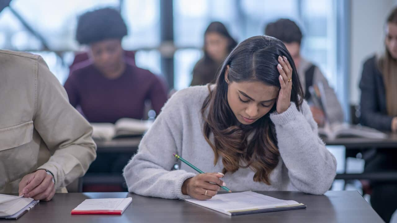 In the classroom, a girl in a light grey sweater is holding her head with one hand, writing with a green pencil on her notebook, looking distressed.