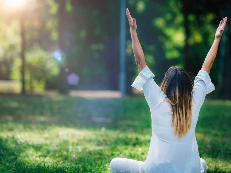 A woman in white is sitting on the grass with her arms raised high, facing away from the camera.